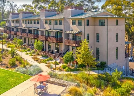 Modern row of three-story townhouses at FiftyOne Baltimore with balconies, surrounded by trees and landscaped greenery; a walking path and a small patio with a red umbrella and chairs are visible in the Crossroads area.