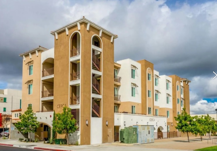 A modern, four-story apartment building with tan and white exterior walls, arched windows, balconies, and surrounding green trees under a partly cloudy sky—ideal for Cerca Student Housing.