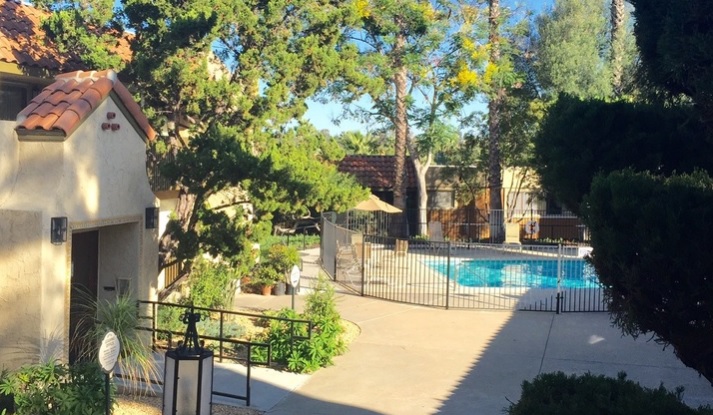 A sunny courtyard at Comanche Hills features a gated outdoor swimming pool, surrounded by trees and shrubbery. Beige stucco buildings with red tile roofs, a walkway, and a black metal fence complete the inviting scene.