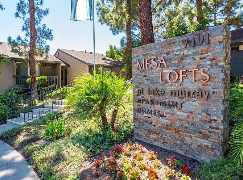 A stone sign reading Mesa Lofts at Lake Murray Apartment Homes stands by a landscaped garden with flowers and greenery, showcasing Elan Mesa Lofts’ luxury lofts in front of a building with tan walls and tall pine trees.