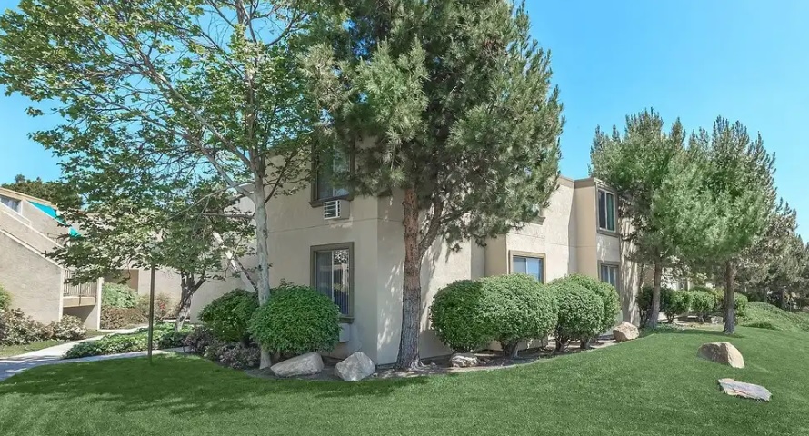 A two-story beige apartment building with large windows is surrounded by green lawns, bushes, rocks, and tall trees under a clear blue sky.