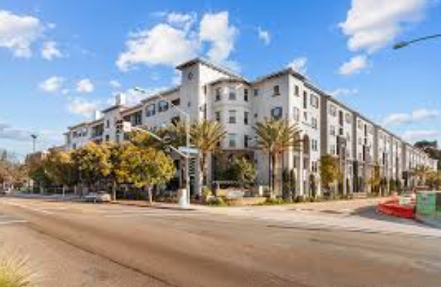 The iconic Alvarado is a modern, multi-story apartment building with white and beige walls, standing on a sunny street corner lined with palm trees and green landscaping under a blue sky with scattered clouds.