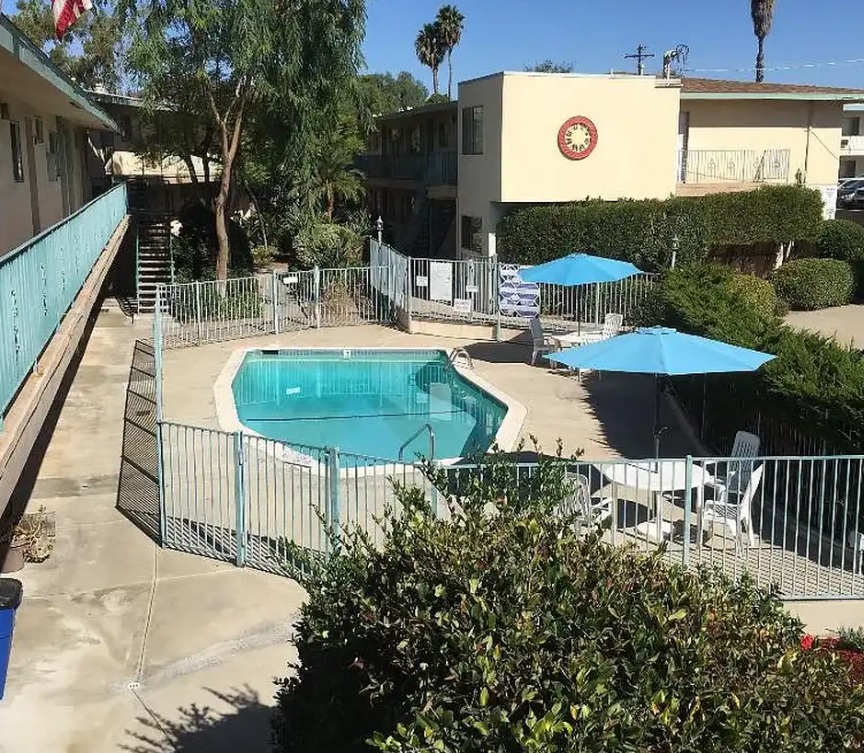 A small outdoor swimming pool surrounded by a metal fence sits in the courtyard of Villa Knolls Apartments, with lounge chairs and blue umbrellas nearby under a sunny sky.