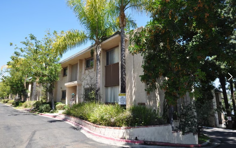 La Cuesta Apartments is a beige two-story building with large windows, surrounded by palm trees and greenery, sitting alongside a sloped street under a clear blue sky.
