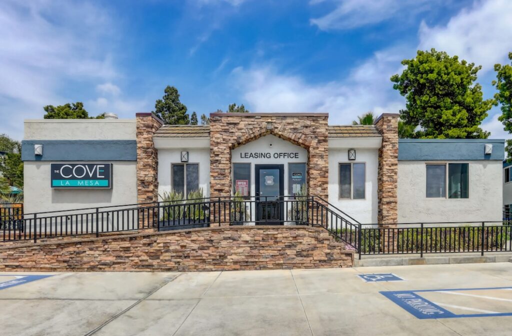 The Cove La Mesa leasing office is housed in a single-story building with a stone and white exterior, glass doors, clear signage, and a ramp. Accessible parking sits in front, while trees and blue sky form the backdrop.