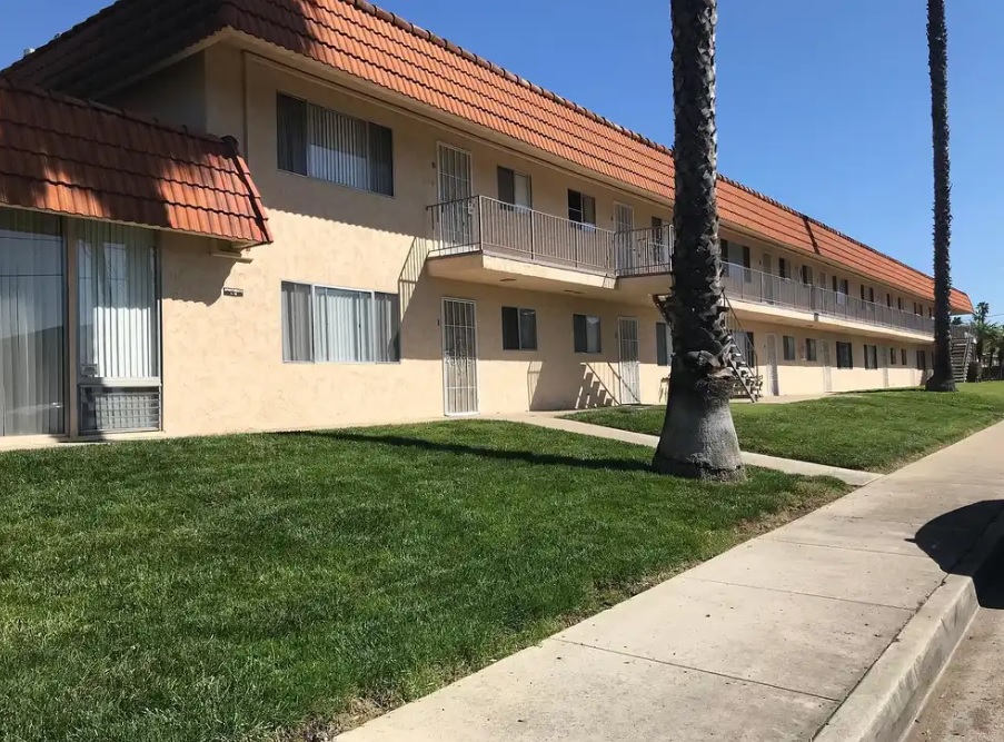 Mohawk Gardens features a two-story beige apartment building with a red-tiled roof, metal railings, and multiple windows. Palm trees line the sidewalk and a well-kept green lawn stretches along the front beneath the clear blue sky.