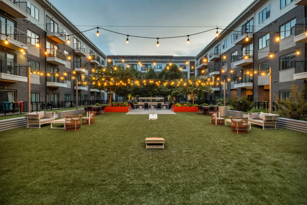 Enjoy the vibrant courtyard at Axis 3700 Plano, featuring green turf, string lights, lounge seating, a cornhole game, lush planters, and modern multi-story buildings with balconies.