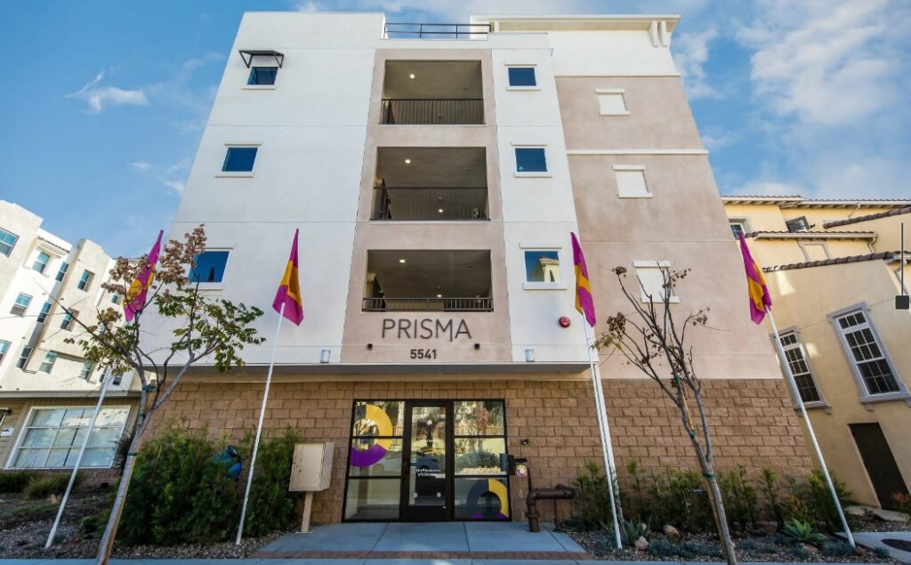 A four-story apartment building with Prisma Student Housing and the number 5541 above the entrance, four colorful flags on poles, a row of small trees, and a partly cloudy sky in the background.