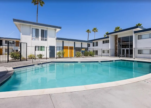 A clear blue swimming pool at State Apartments San Diego is surrounded by a concrete deck and black metal fence, with two white, two-story apartment buildings and several palm trees in the background under a bright blue sky.