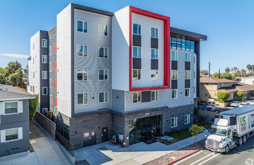 Modern five-story apartment building with a white, gray, and black facade and a large red rectangular accent around upper windows, located next to residential homes; a white truck is parked in front.