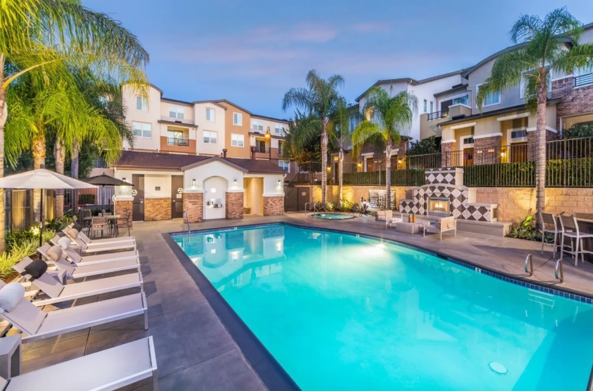 A modern outdoor swimming pool surrounded by lounge chairs, palm trees, and apartment buildings at sunset, with a fire pit and decorative wall in the background.