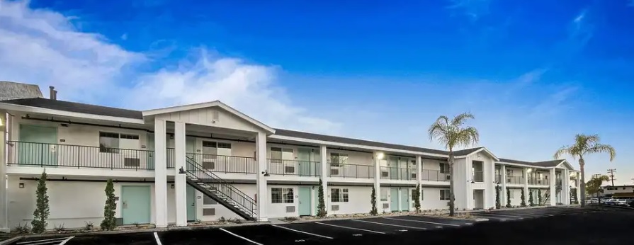 A two-story motel with white exterior walls, exterior staircases, palm trees in front, and an empty parking lot under a bright blue sky.