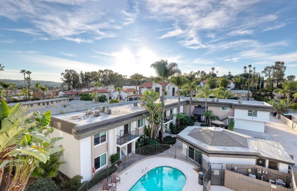 Aerial view of The Village at La Mesa, a sunny residential neighborhood featuring a swimming pool, modern low-rise buildings, palm trees, and a clear blue sky with scattered clouds.