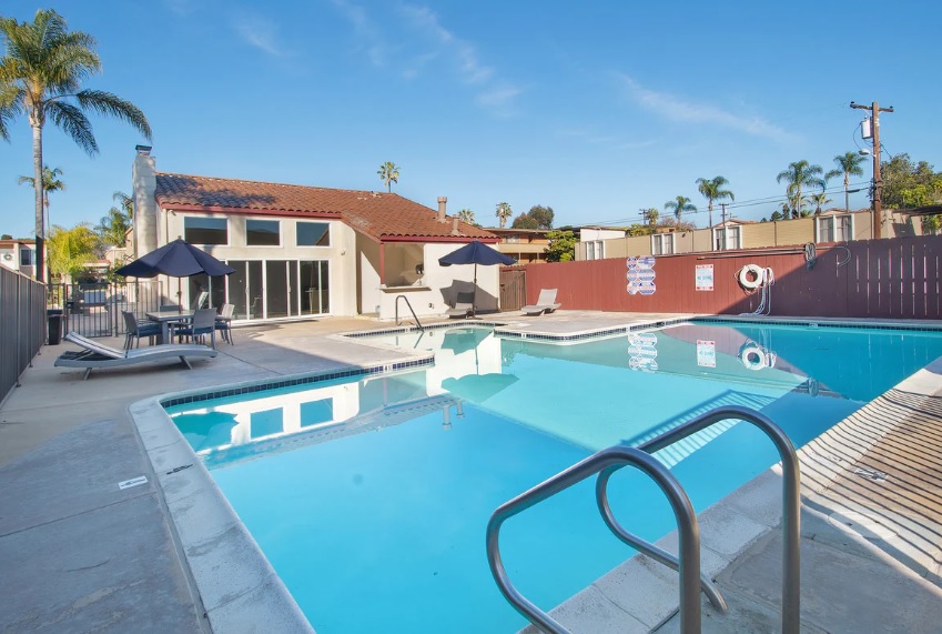 Outdoor swimming pool with clear blue water, lounge chairs, and umbrellas beside the Tierra Del Rey building with large windows. Palm trees and a clear blue sky complete this relaxing retreat.