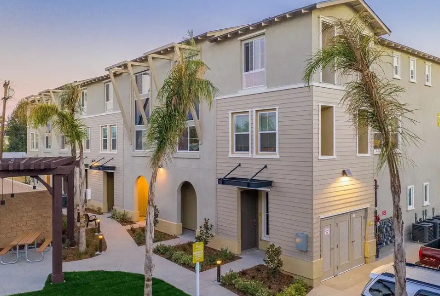 Modern three-story Trio Townhomes feature a beige and light gray exterior, large windows, and palm trees along the walkway. A picnic table sits on the patio to the left, with lush green landscaping welcoming you at the entrance.