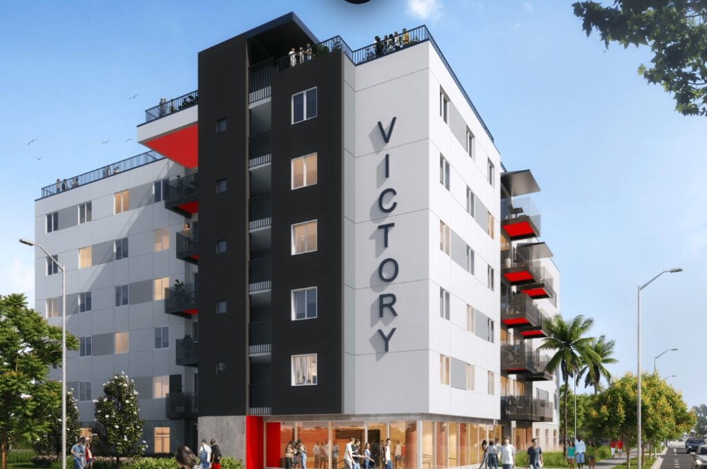 A modern six-story apartment building, Victory on Paseo, displays VICTORY vertically on its facade. Balconies and large windows overlook people strolling outside on a sunny day, surrounded by palm trees and lush greenery.