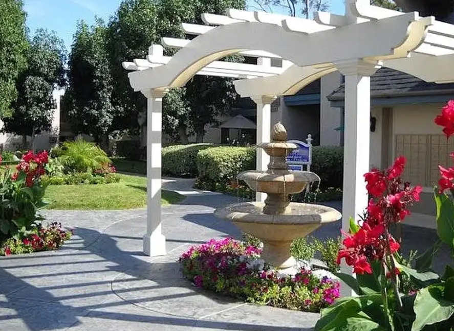 A stone fountain sits beneath a white pergola in the landscaped garden at Village Apartments, surrounded by blooming red flowers, green bushes, and trees on a sunny day. Paths and buildings are visible in the background.