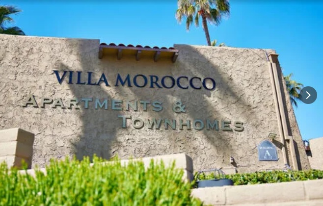 A beige stucco building displays a “VILLA MOROCCO APARTMENTS & TOWNHOMES” sign in blue letters, with palm trees and a bright blue sky, capturing the serene charm of Villa Morocco.