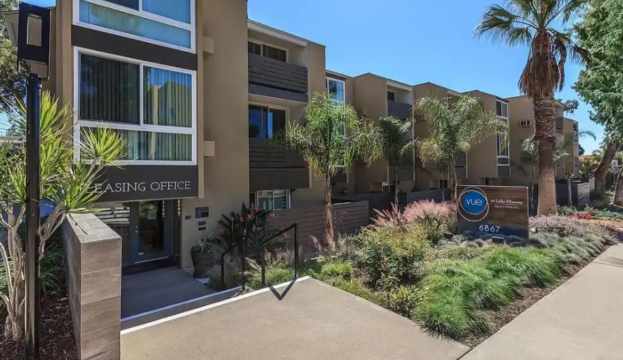 Modern apartment complex with beige buildings, balconies, large windows, and lush landscaping. A sign near the sidewalk reads Leasing Office and includes the address 6867. Palm trees and plants line the walkway at Vue at Lake Murray.