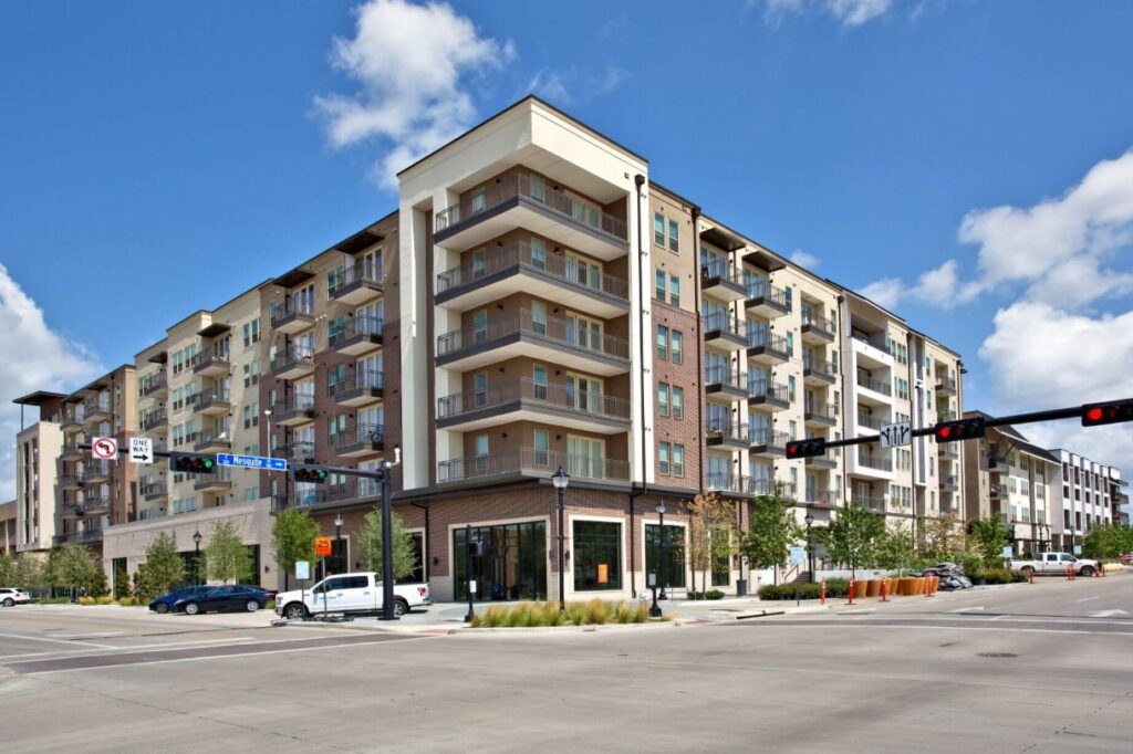 A modern six-story apartment building, 101 Center Arlington, stands on a street corner with balconies, large windows, and ground-floor shops. Traffic lights, cars, and greenery line the intersection under a partly cloudy sky.