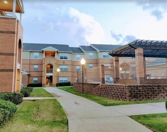 A three-story brick apartment building at Arbor Oaks Arlington features lit street lamps, a gazebo with a pergola roof, and a paved walkway surrounded by grass and shrubs in the foreground at dusk.