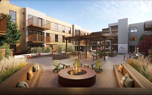 A modern apartment courtyard with a central fire pit, wooden seating with pillows, patio tables and chairs, greenery, and three-story buildings with balconies under a blue sky.