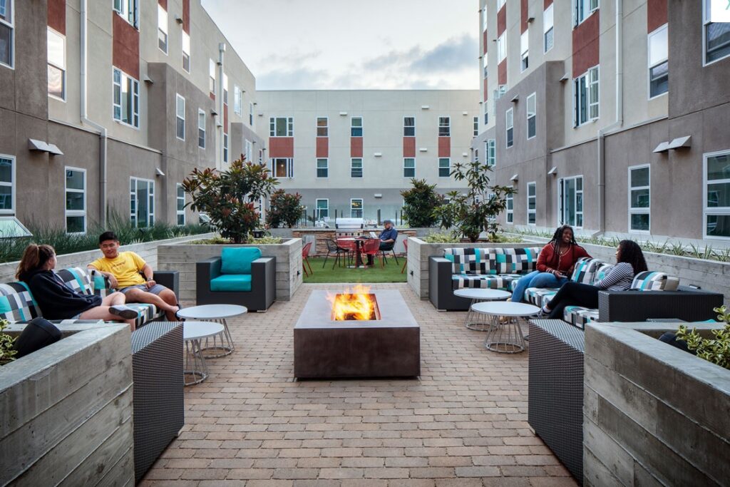 Residents at M at College San Diego sit and relax on cushioned outdoor furniture around a rectangular fire pit in a modern apartment courtyard, surrounded by greenery, tables, and multi-story buildings in the background.