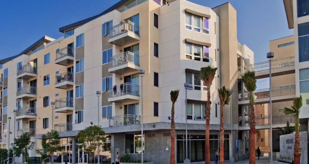 Modern five-story apartment building with balconies, large windows, and tan exterior. Palm trees line the sidewalk at Paseo Place San Diego, and the building features a glass entrance at street level.